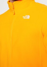 Chaqueta de forro polar amarilla brillante con cremallera frontal y el logo "The North Face" bordado en el pecho.