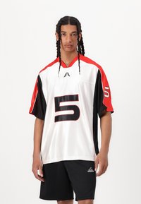 Camiseta atlética blanca con acentos rojos y negros, con un gran número 5 negro, corte holgado, mangas cortas y textura de malla. Pantalones cortos negros.