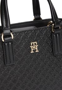 Μαύρη δερμάτινη τσάντα χειρός με ανάγλυφο γεωμετρικό σχέδιο, χρυσαφί λογότυπο "TH" και διπλές λαβές με χρυσούς κρίκους.