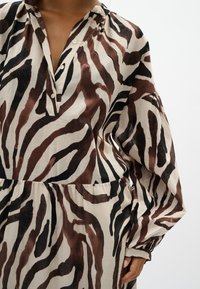 Gros plan d'une personne portant une robe beige à manches longues avec des rayures zèbre abstraites marron et noires et un col en V boutonné.