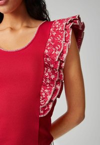 Débardeur rouge à côtes avec des manches volantées à motif floral. Les détails notables incluent une bordure contrastante le long de l'encolure et des bords de manches.