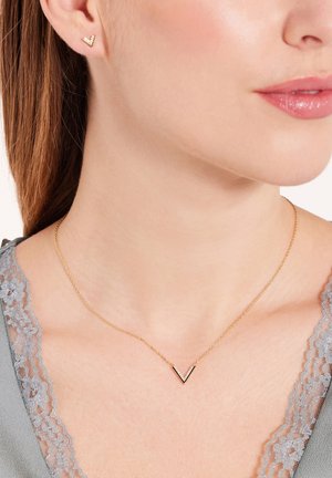 Goldkette mit einem V-förmigen Anhänger, der mit kleinen klaren Steinen besetzt ist, kombiniert mit passenden goldenen Stecker Ohrringen, getragen über einem grauen Spitzenoberteil.