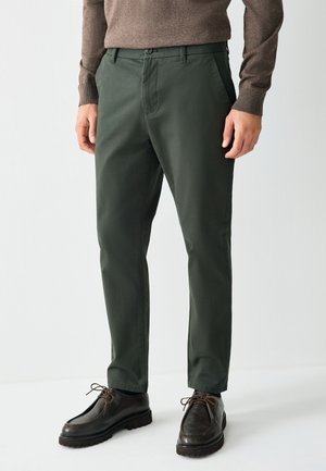 Hombre con pantalones ajustados verde oscuro, suéter marrón de manga larga y zapatos de cuero marrón oscuro con cordones, de pie sobre fondo blanco.