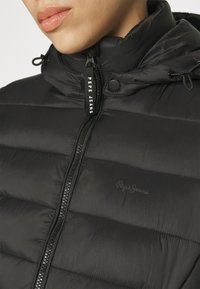 Gros plan d'une veste matelassée noire avec une fermeture éclair et le logo "Pepe Jeans" sur la poitrine et le curseur de la fermeture éclair, portée par une personne.