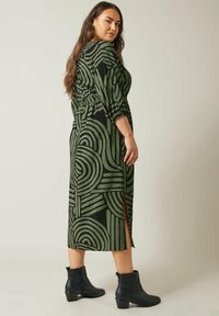 Langes schwarzes Kleid mit grünem abstraktem Muster, mit betonter Taille und seitlichen Schlitzen. Kombiniert mit schwarzen Knöchelstiefeln.