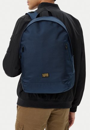 Muž v černé bundě a hnědých kalhotách s tmavě modrým batohem G-Star Originals na zádech.