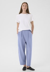 Bijela majica s kratkim rukavima u kombinaciji s svijetloplavim plisiranim hlačama; model nosi crne slip-on cipele; glatka tkanina, opušteni kroj.
