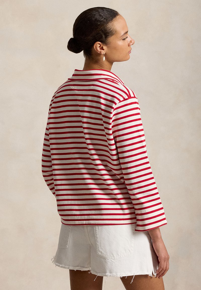 Frau mit Dutt, die ein rot-weiß gestreiftes Langarmshirt und weiße Jeansshorts trägt, steht vor einem einfarbigen beigen Hintergrund.