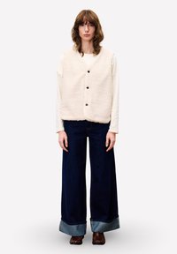 Junge Frau mit schulterlangem Haar, die eine weiße, geknöpfte Fleeceweste, ein langärmliges Hemd, weite dunkle Jeans mit Umschlägen und braune Schuhe trägt.