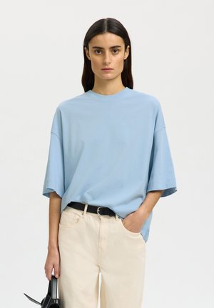Vrouw met steil donker haar, draag een losse lichtblauwe blouse en crèmekleurige broek, houdt een zwarte handtas vast met één hand in de zak.