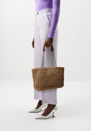 Borsa a mano in paglia intrecciata con dettagli in pelle marrone, tracolle a catena argentata e forma rettangolare, tenuta da una persona che indossa pantaloni viola chiaro.