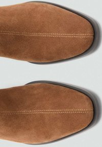 Un paio di stivali marroni in suede con cuciture visibili al centro e suole nere su uno sfondo grigio chiaro.
