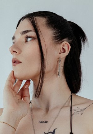 Silver dangling earrings with intricate chain design, worn by a person with dark hair, smooth skin, and visible tattoos. Light background.