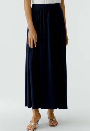 Frau trägt einen langen dunkelblauen Rock kombiniert mit silbernen Sandalen mit Absatz, steht vor einem schlichten, hellen Hintergrund.