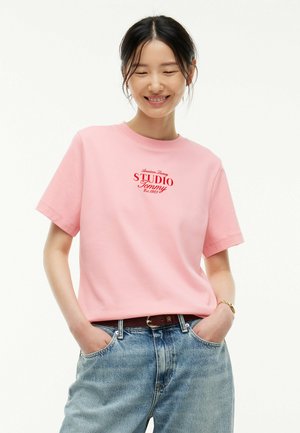 Jeune femme souriante, les mains dans les poches, portant un t-shirt rose avec un texte rouge et un jean bleu sur un fond blanc uni.