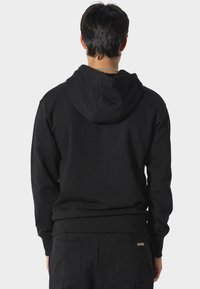 Hombre con sudadera negra y pantalones negros, de espaldas a la cámara, contra un fondo gris claro liso.