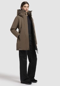 Hnedá izolovaná parka s vysokým golierom, zipsom a sťahovacími šnúrkami. Nosená cez tmavé nohavice, s bočnými vreckami a elegantným dizajnom.