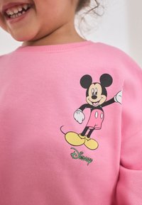 Roosa dressipluus, millel on musta ja kollase trükiga Mickey Mouse ning rohelise "Disney" logoga, valmistatud pehmest, struktuursed kangast.