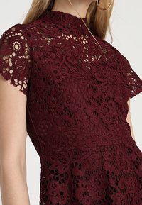 Burgundy spetsklänning med korta ärmar, hög halsringning, blommönster och intrikata utskärningar som visar en strukturerad tygdesign.
