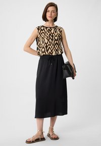 Mouwloos topje met een zwart en beige abstract patroon, gecombineerd met een zwarte drawstring rok. Geaccentueerd met gouden sandaaltjes met banden en een kleine zwarte tas.