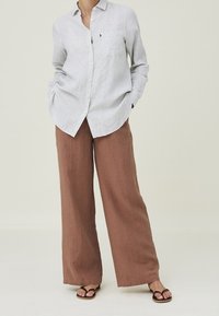 Chemise boutonnée en lin gris clair avec poche, associée à un pantalon large en lin marron. Le modèle porte des sandales noires, les mains dans les poches.