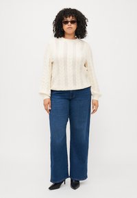 Cream cable-knit sweater with a round neckline, paired with wide-leg blue jeans and black pointed-toe heels, accessorized with sunglasses.