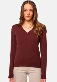 Pull à col en V bordeaux avec des manches longues, fabriqué à partir d'un matériau doux. Poignets et ourlet côtelés. Porté sur un pantalon clair.