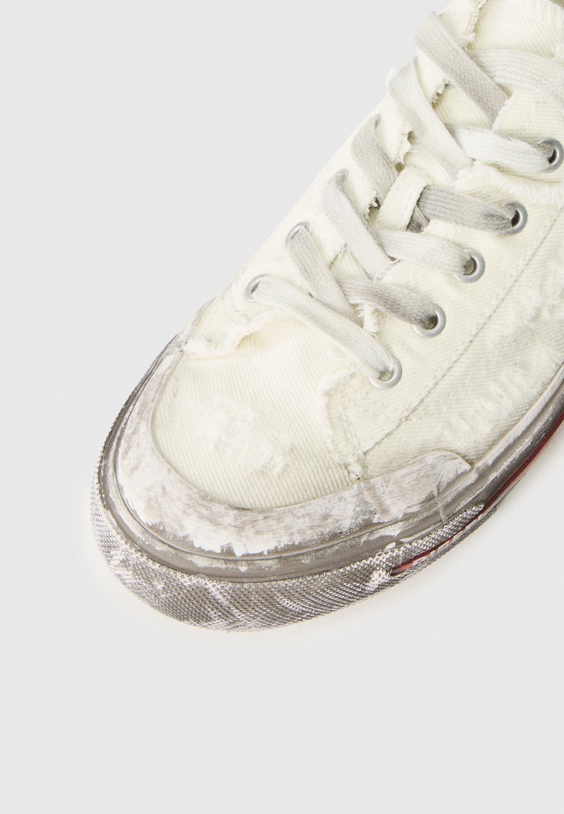 Zapatilla de lona blanca con bordes deshilachados y desgaste visible. Suela de goma plateada con acento texturizado, con cordones gris claro y ojales de metal.