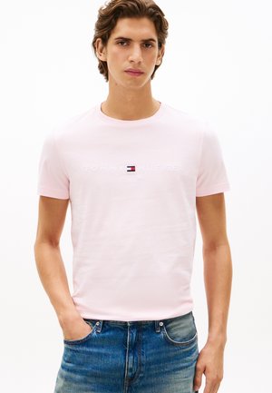 Tânăr cu păr de lungime medie purtând un tricou roz deschis Tommy Hilfiger și blugi albaștri, stând cu o mână în buzunar.