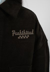 Dunkelbraune Cordjacke mit einer Metallnadel, die das Wort "Verboten" über einem Schachbrettmuster auf der Brust zeigt.
