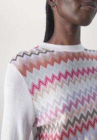 Personne à la peau foncée aux cheveux tressés porte un pull blanc avec des motifs zigzag multicolores et une boucle d’oreille en or.