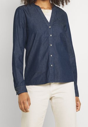 Femme portant une chemise en denim bleu foncé boutonnée à manches longues et un pantalon beige clair, debout devant un fond uni.