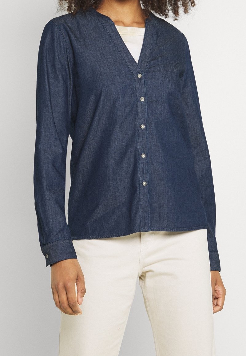 Femme portant une chemise en denim bleu foncé boutonnée à manches longues et un pantalon beige clair, debout devant un fond uni.