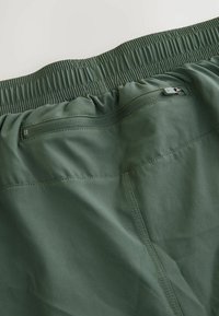 Grønne shorts med elastisk talje, der har en lynlåslomme og en glat, let stofstruktur. Syninger synlige langs sømmene.