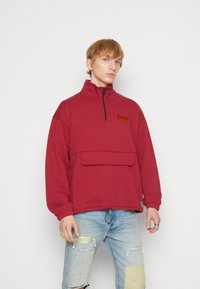 Felpa rossa con collo alto, mezza zip e taschino frontale; presenta un logo in ricamo marrone testurizzato; vestibilità comoda, in misto cotone.