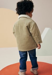 Giacca di velluto a coste verde oliva con colletto in sherpa bianco, abbinata a jeans blu e scarpe beige, vista da dietro.