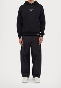 Czarna bluza z kapturem z przednią kieszenią i logo, zestawiona z luźnymi czarnymi spodniami typu cargo z kieszeniami po bokach oraz białymi sneakersami.