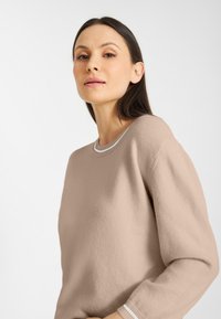 Vrouw met lang donker haar in een beige trui met witgestreepte manchetten, die iets naar boven kijkt tegen een effen lichte achtergrond.