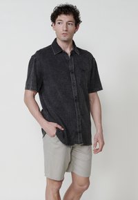 Camisa negra de manga corta con textura, bolsillo frontal, botones en la parte delantera y pantalones cortos ligeros en color beige. Patrón sutil, ajuste relajado.