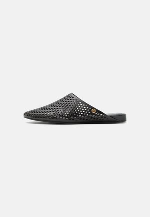 Μαύρο mule slip-on με ανοιχτές διαμαντένιες οπές και μικρό στρογγυλό χρυσό λογότυπο στο πλάι, επίπεδη σόλα, μινιμαλιστικός σχεδιασμός.