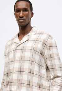 Károvaná košeľa v béžových, oranžových a čiernych odtieňoch. Má vypilovaný golier, zapínanie na gombíky a mäkkú látku s voľným strihom.