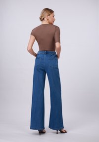 Bodysuit marrone a maniche corte abbinato a jeans blu a vita alta e gamba larga, con tasche posteriori e una vestibilità rilassata, su uno sfondo neutro.