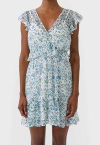 Femme portant une robe blanche à motif floral bleu, avec des manches courtes volantées, un décolleté en V et un ourlet volanté, se tenant contre un fond uni.