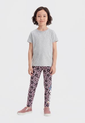 Enfant aux cheveux courts et foncés debout, portant un t-shirt gris clair, des leggings roses avec un imprimé floral noir et un détail de dessin animé, ainsi que des chaussures roses sans lacets.