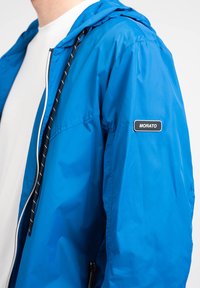 Blå letvægtsjakke med hætte, der har lynlåslukning og snoretræk. Sort accent med teksten "MORATO" på venstre ærme.