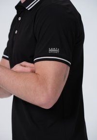 Camisa polo negra con rayas blancas contrastantes en el collar y las mangas. Presenta un pequeño logo "LUHTA SPORT" en la manga izquierda.