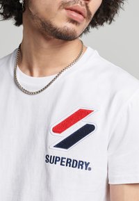 T-shirt bianco in cotone con un logo ricamato multicolore con accenti rossi, blu e bianchi. Collana a catena d'argento visibile.