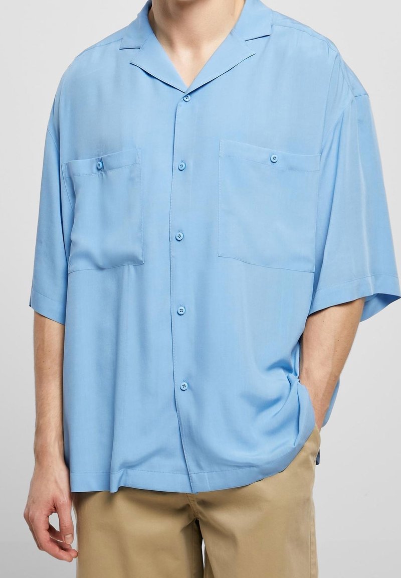 Chemise bleu clair à manches courtes avec boutons et deux poches poitrine portée sur un pantalon beige, une main dans la poche, sur une personne debout.