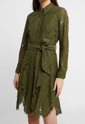 Koronkowa sukienka w oliwkowo-zielonym kolorze z długimi rękawami, kołnierzem, wiązanym paskiem w talii oraz wykończeniem w falbanki, noszona przez osobę na jednolitym tle.