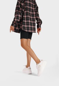 Sneakers bianchi con una finitura lucida e logo, abbinati a pantaloni in denim neri e una camicia oversized a quadri rosa.